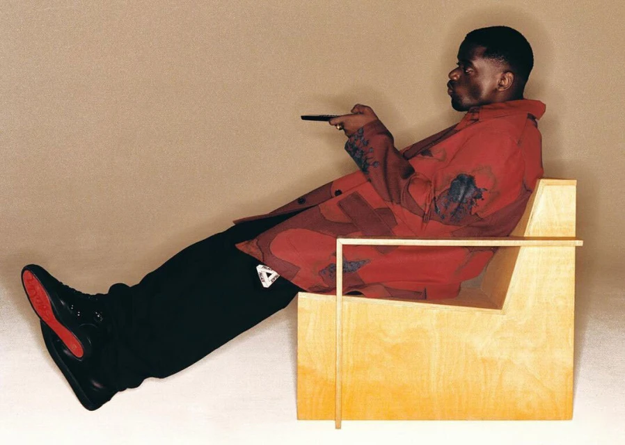 An adult Black man lounges in a modern wood chair, holding a remote, wearing black pants, matching boots, and a patterned red jacket in studio light.