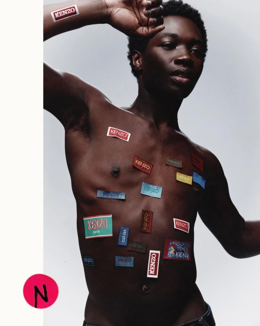 Young adult poses shirtless in a studio setting covered in Kenzo brand stickers. Fashion forward image with unique styling.