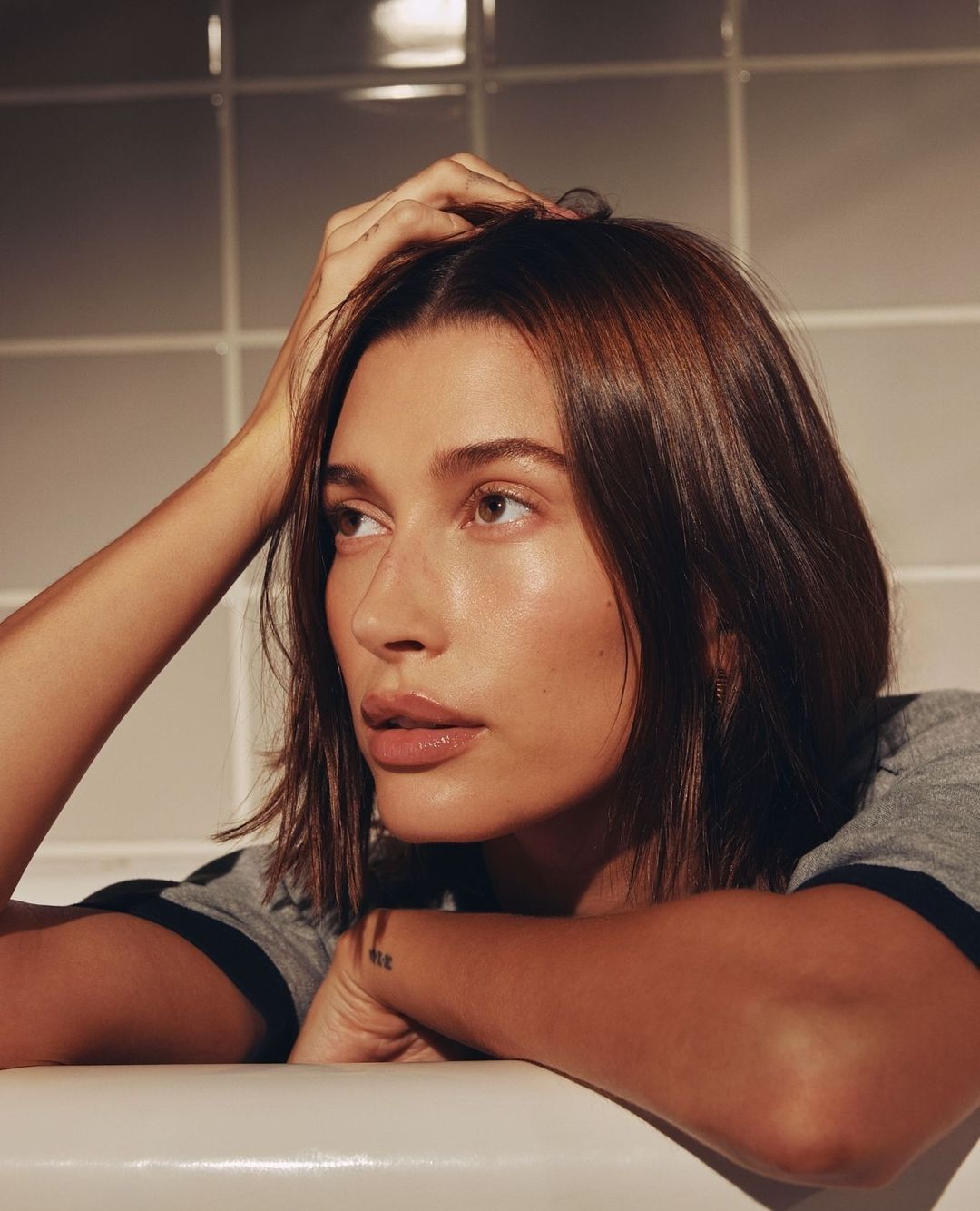 Close-up portrait of a brunette adult woman with hand in hair, wearing a gray shirt, resting her arms on a white bathtub edge.