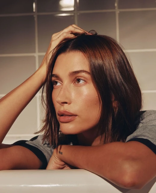 Close-up portrait of a brunette adult woman with hand in hair, wearing a gray shirt, resting her arms on a white bathtub edge.