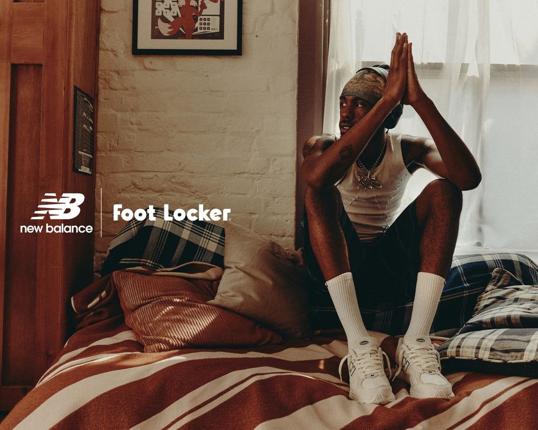 A young adult male wearing a bandana and athletic shoes poses on a striped bed, near a window, for a New Balance and Foot Locker ad.