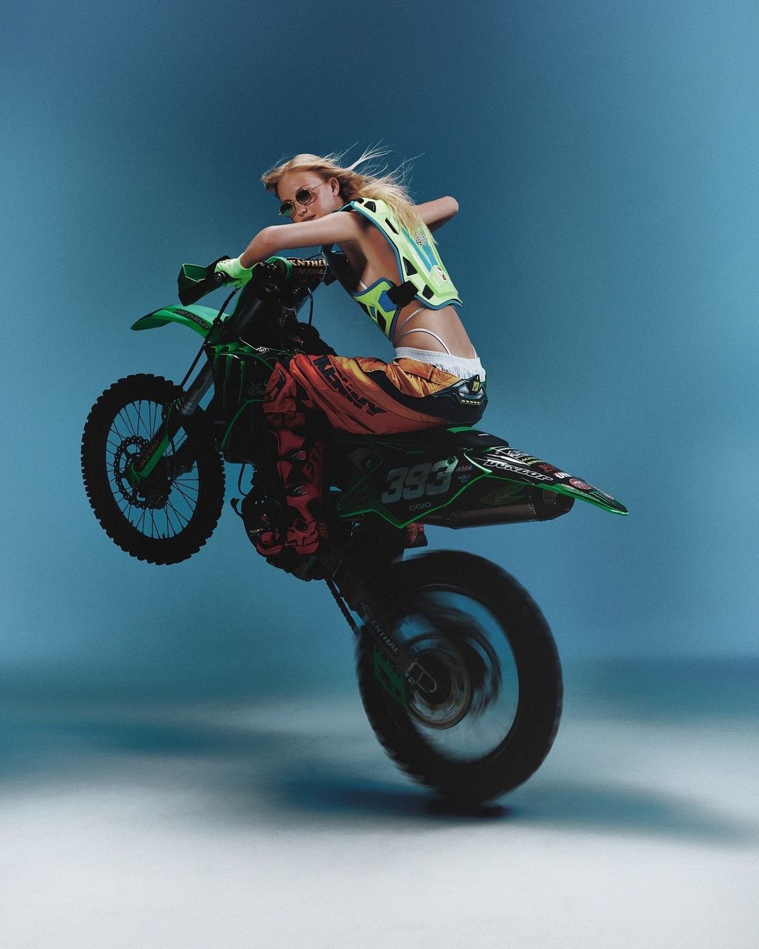 A blonde woman wearing safety gear performs a wheelie on a green dirt bike against a light blue backdrop in an action studio shot.