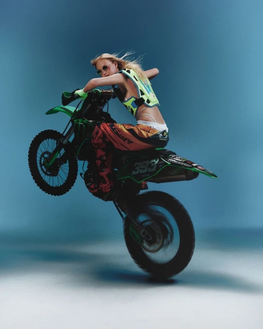 A blonde woman wearing safety gear performs a wheelie on a green dirt bike against a light blue backdrop in an action studio shot.