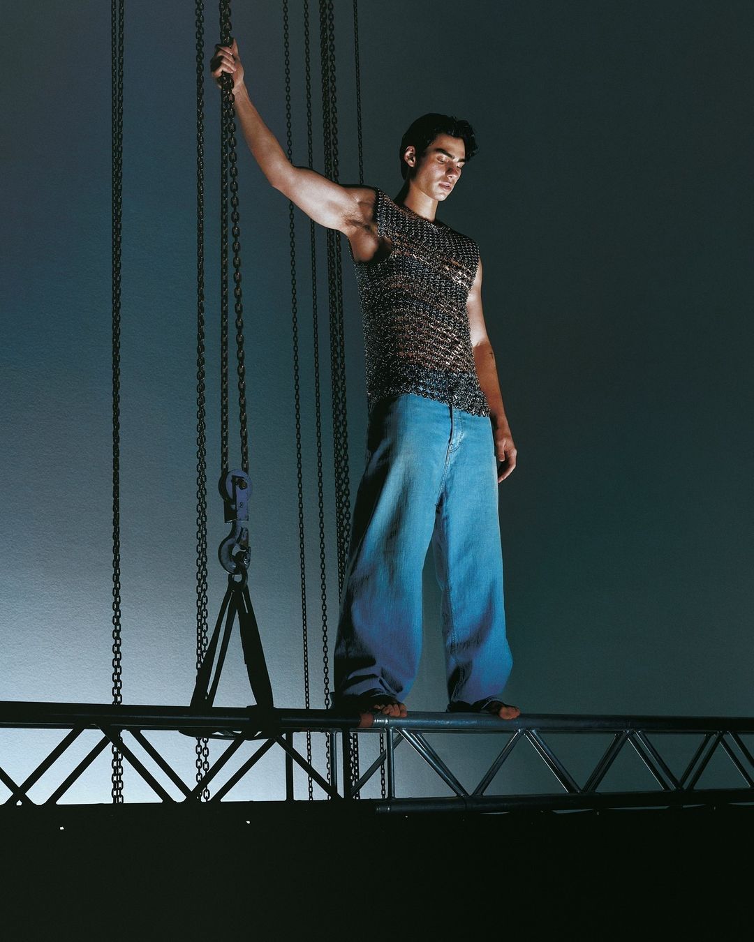 A male model stands on a metal structure, wearing a textured top and jeans, with hanging chains in a studio setting.