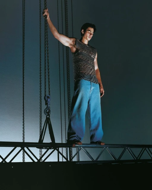 A male model stands on a metal structure, wearing a textured top and jeans, with hanging chains in a studio setting.