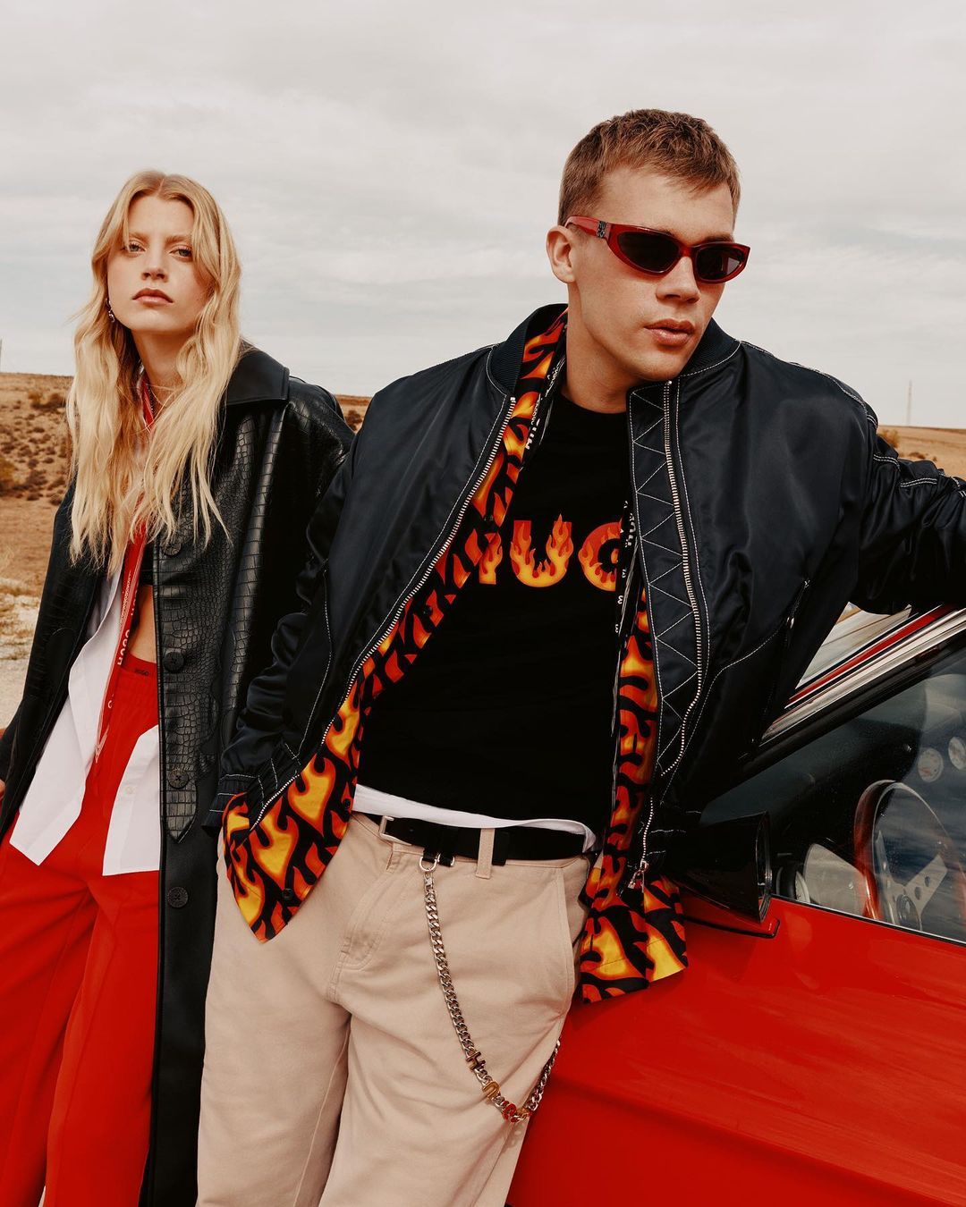 Two fashion models pose next to a red car on a cloudy day in a rural setting, wearing a bomber jacket, leather coat and sunglasses.