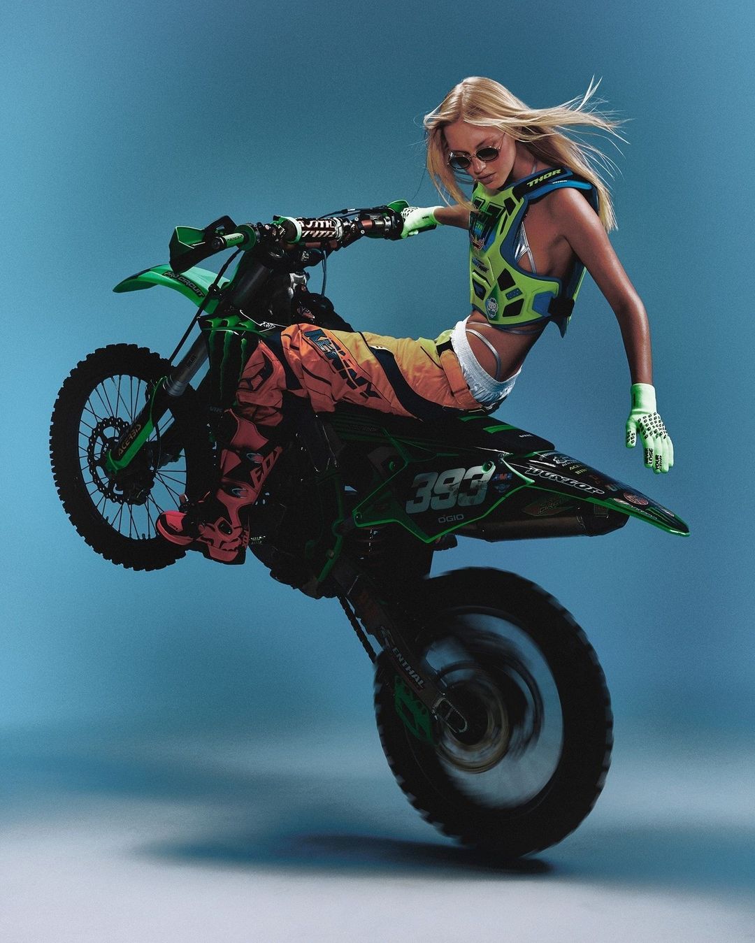 Woman performing a wheelie on a green and black dirt bike against a blue backdrop in an action shot with full gear and protective vest.