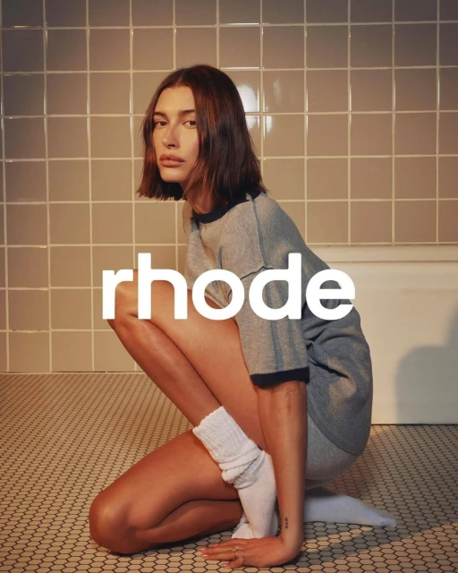 Adult female in t-shirt and shorts poses kneeling in bathroom with white block letters 'rhode' in foreground on tiled floor and walls.
