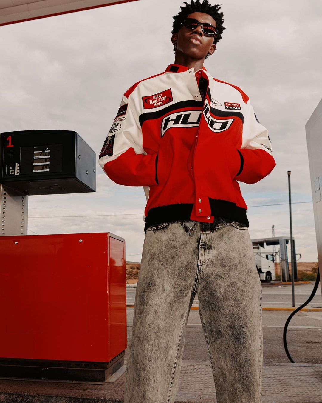 Man with sunglasses and stylish red and white racing jacket poses in front of a gas pump at an outdoor gas station.