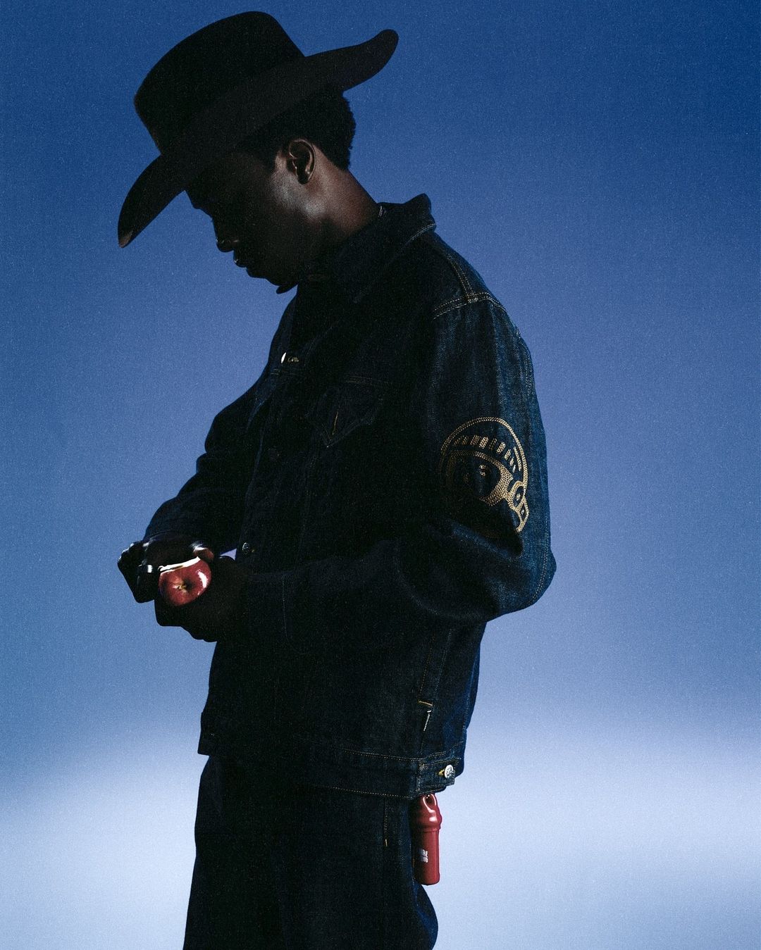 Stylish African-American male model wearing a denim jacket and cowboy hat poses with an apple against a blue backdrop in a studio shot.