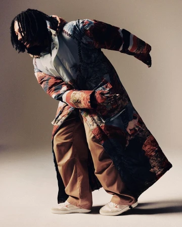 Man wearing a long printed coat and tan trousers leaning forward in a studio