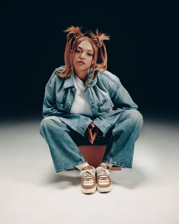 Woman in denim jacket and matching jeans with pigtails crouching against a dark gradient backdrop