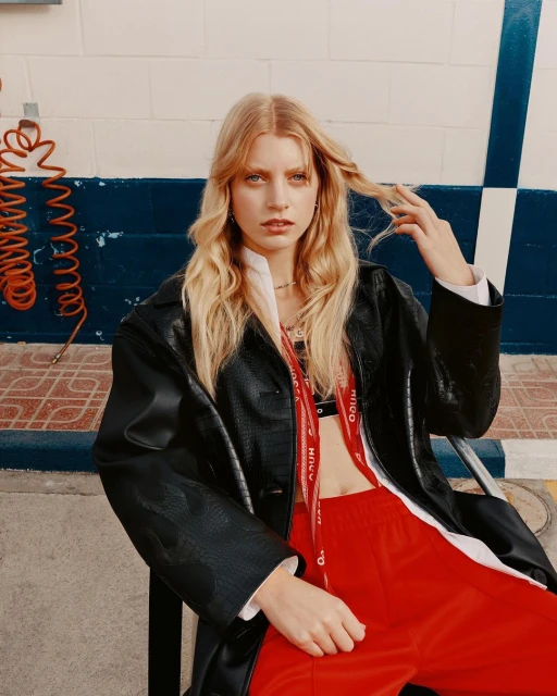 A blonde model in a black jacket and red pants poses against a white wall. The model is seated and has her hand in her hair.
