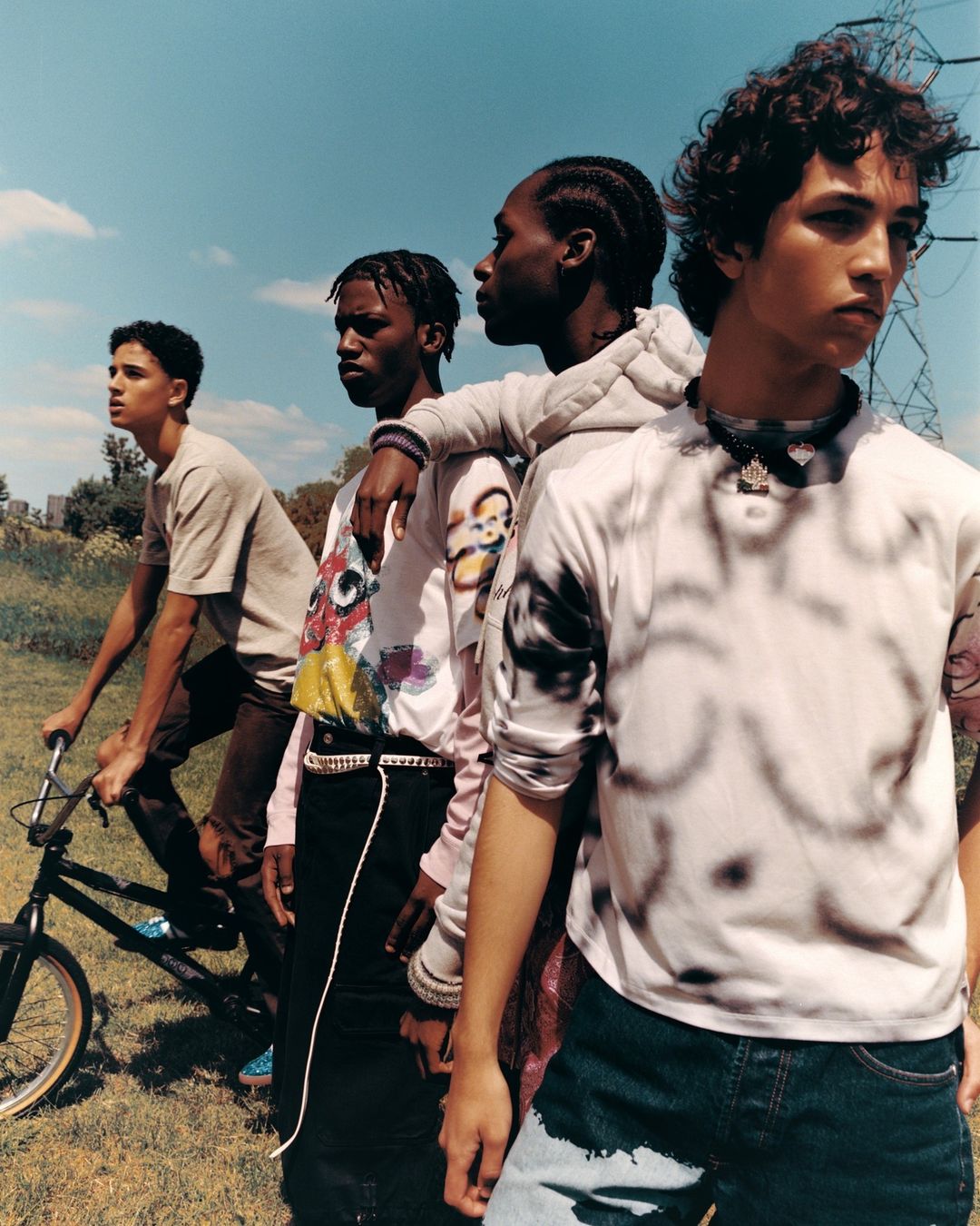 Four young men outdoors, one on a bicycle, dressed in casual streetwear, posing against a blue sky, evoking a casual and youthful mood.