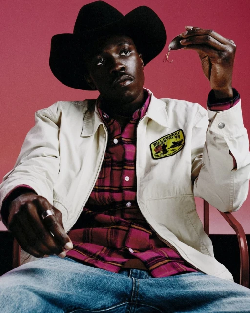 A black male model in a cowboy hat and jacket holds a fishing lure while posing indoors against a red background in a medium shot.