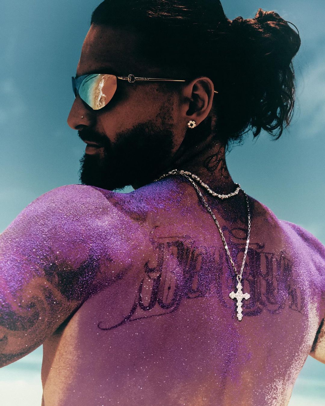Profile portrait of a man with glitter on his back, sunglasses, and a hair bun, posing against a blue sky backdrop.