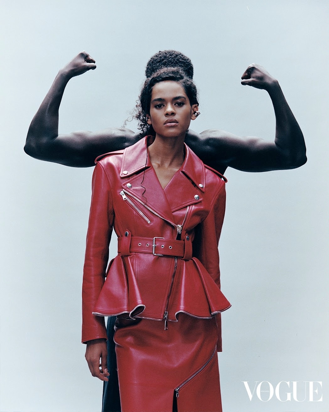 Editorial shot of an African woman in a red leather jacket and skirt, posing in front of a strong man's arms in a studio setting.