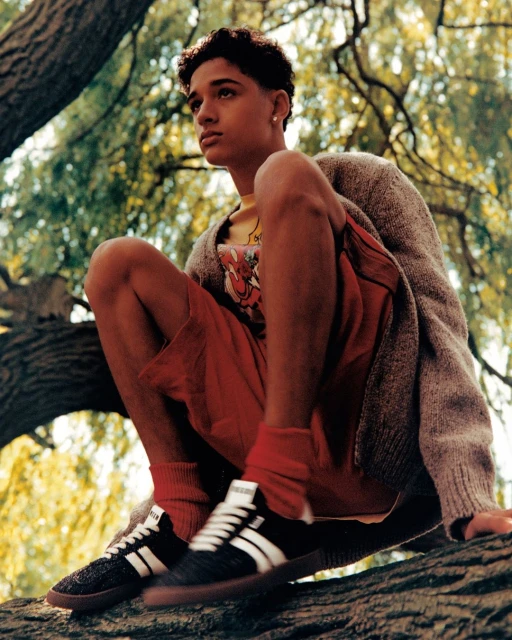 Teenager sits on a tree branch wearing a cardigan, t-shirt, shorts, socks, and sneakers in a green outdoor setting with trees.