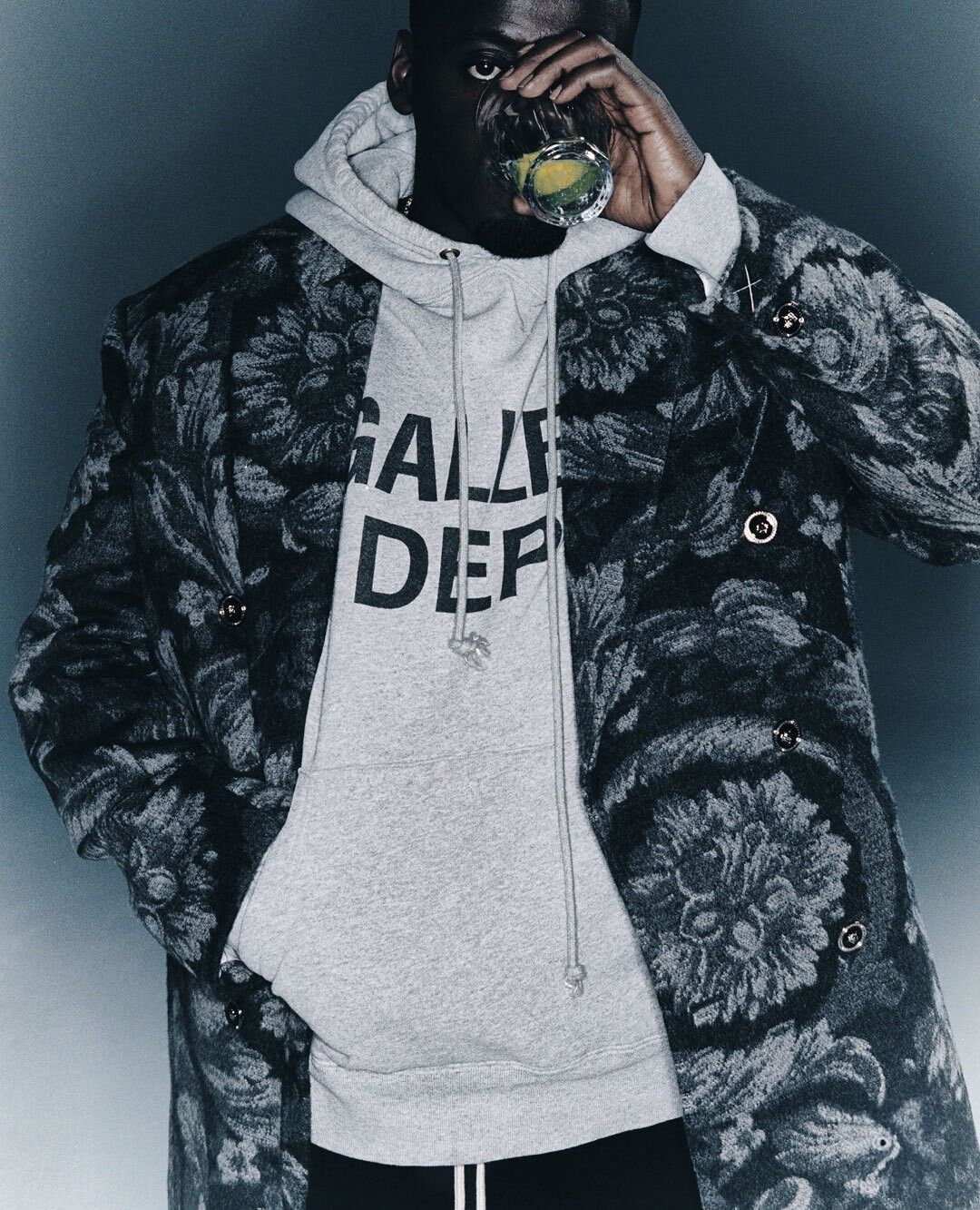 Adult Black man wearing Gallery Dept. hoodie and patterned coat, holding a glass and drinking, against a plain grey backdrop.