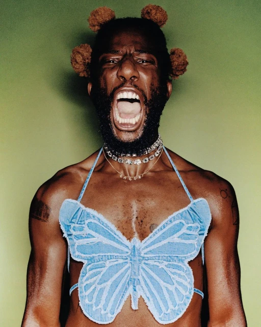 Striking studio portrait of a Black male model with pom-pom hair, wearing a light blue denim butterfly top, and multiple chain necklaces.