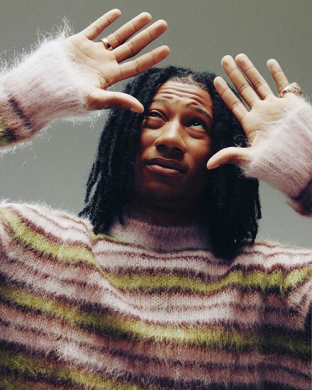 Portrait of a Black male with dreadlocks wearing a striped fuzzy sweater with his hands up, shot from a low angle against a plain background.