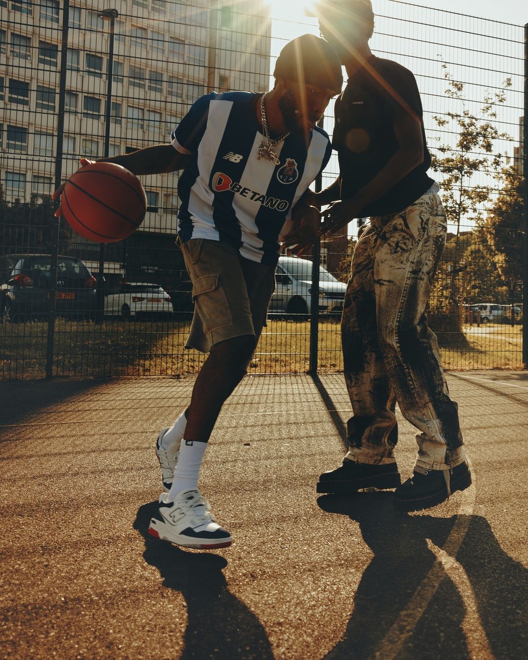 Two young adult men play basketball on an outdoor court; one dribbles with a basketball, wearing a sports jersey and white sneakers.
