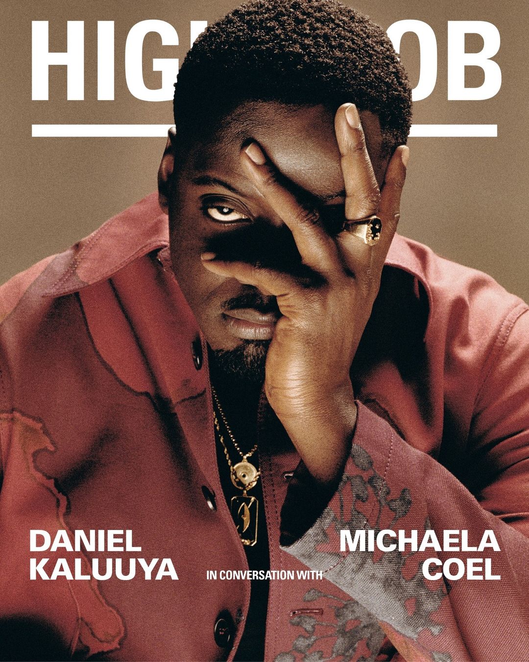 Portrait of Daniel Kaluuya in a red jacket with his hand near his face, a gold necklace, and a ring, against a tan backdrop.