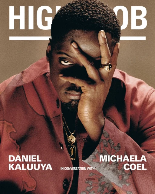 Portrait of Daniel Kaluuya in a red jacket with his hand near his face, a gold necklace, and a ring, against a tan backdrop.