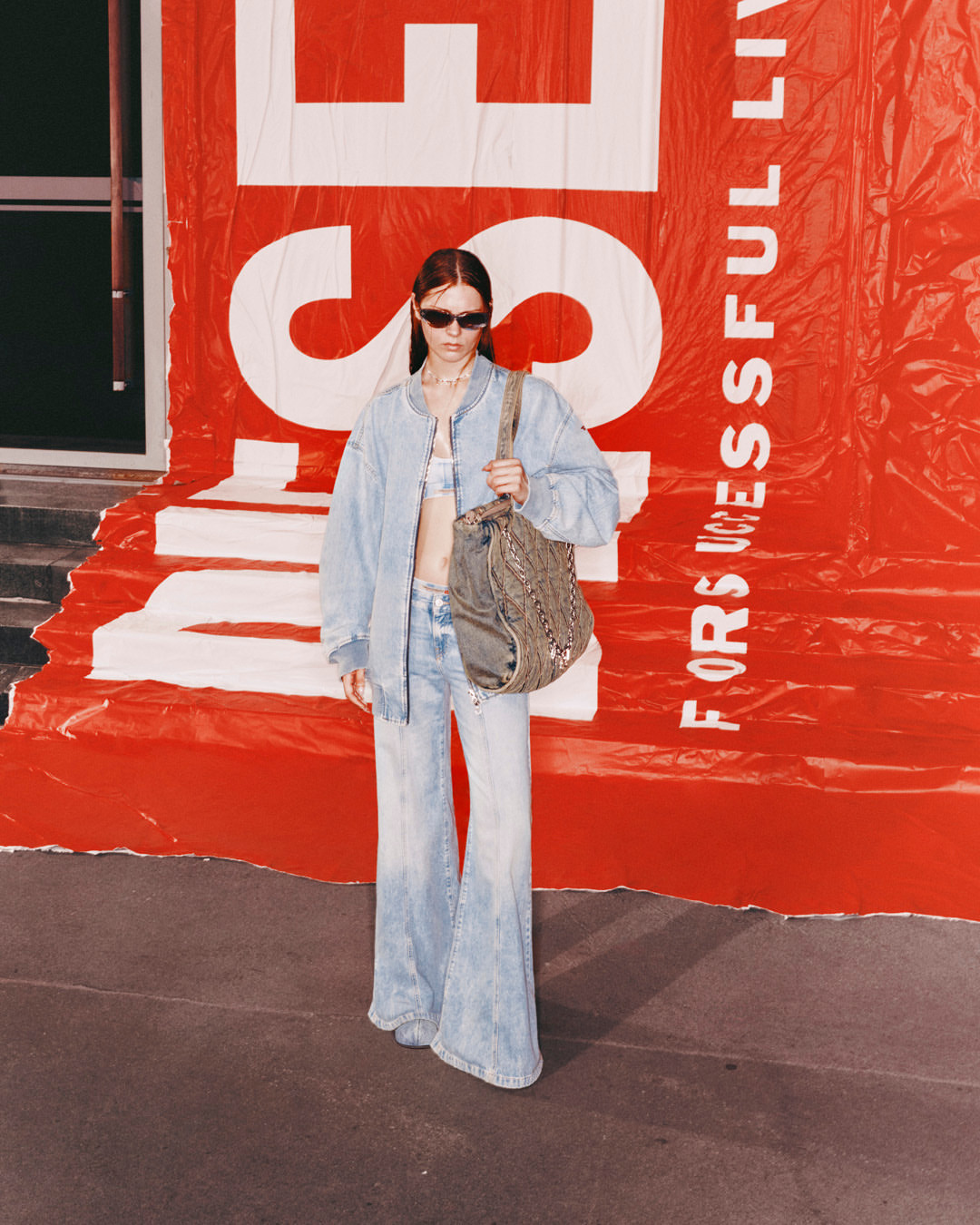 A stylish young adult female in denim outfit carrying a handbag, posing in an urban setting in front of a large red advertisement.