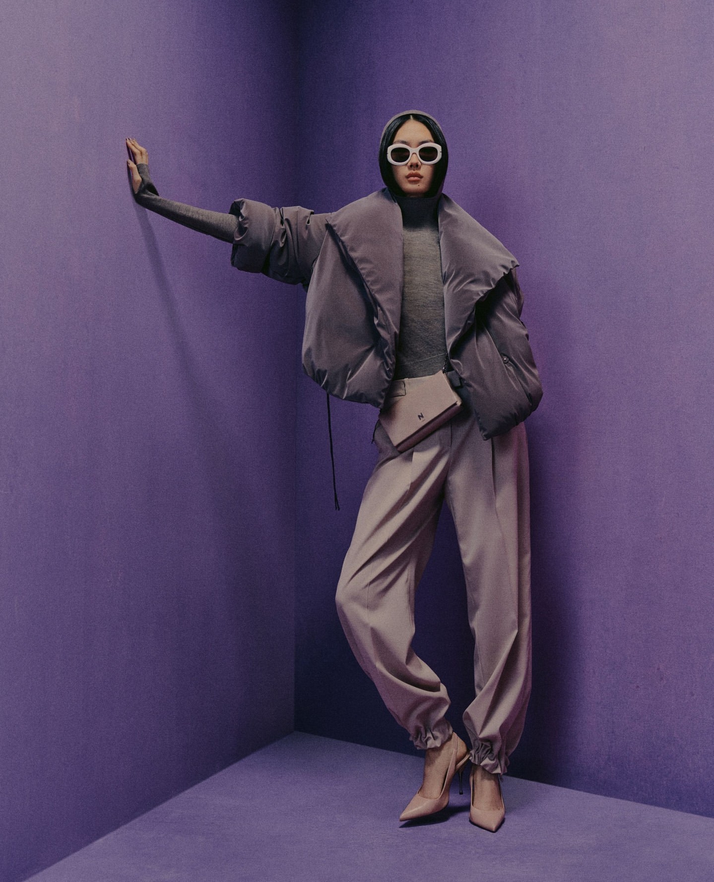 Woman in lavender puffer jacket, turtleneck, pants, heels, and sunglasses poses in a lavender corner setting for a fashion shot.