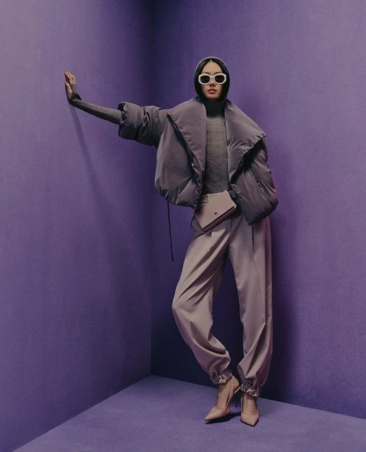 Woman in lavender puffer jacket, turtleneck, pants, heels, and sunglasses poses in a lavender corner setting for a fashion shot.