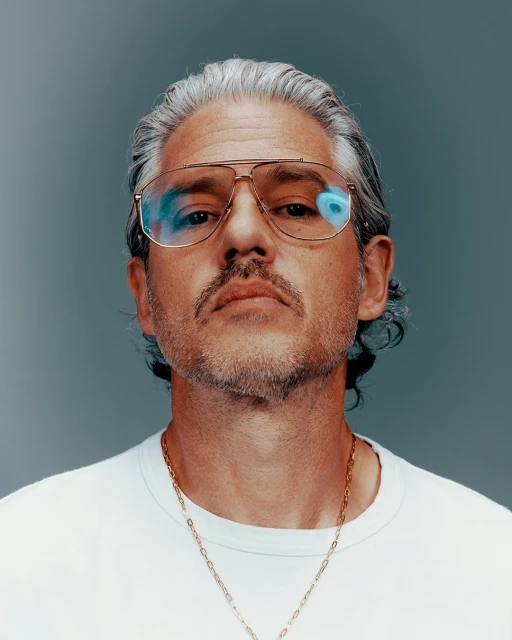 Close-up portrait of a middle-aged adult male with gray hair wearing aviator glasses and a white t-shirt against a grey background.