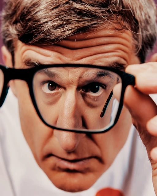 Close-up portrait of an adult male looking through eyeglasses, with a light complexion, brown hair, and a light purple backdrop.