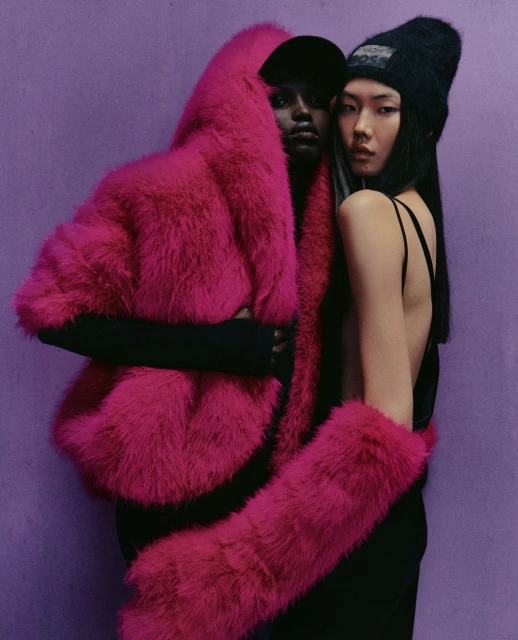 Two women pose against a light purple background, one in a hot pink faux fur coat and the other in a black top and beanie.