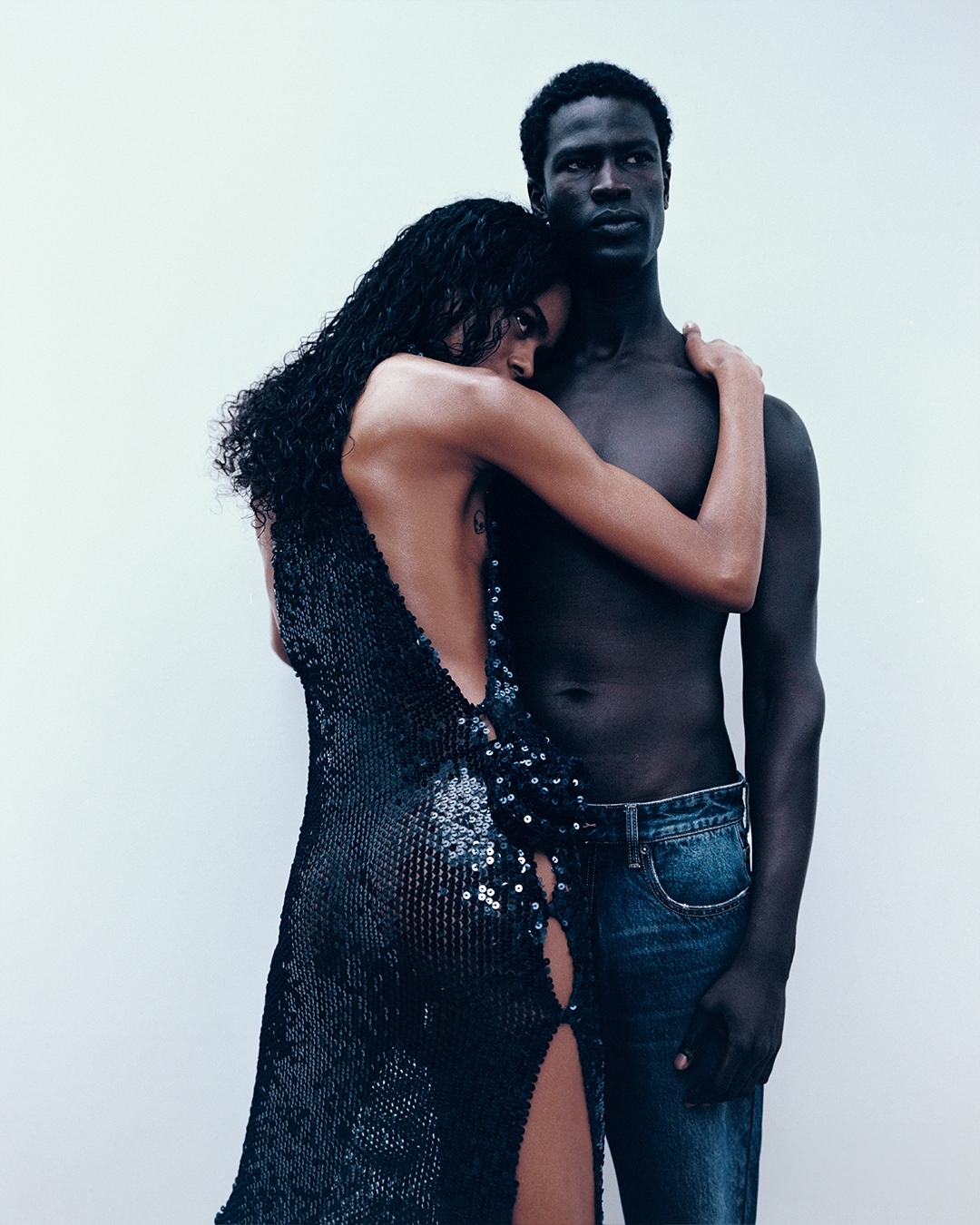 Striking studio shot of an embracing interracial couple: a Black man with bare torso and a woman in a black sequined dress.