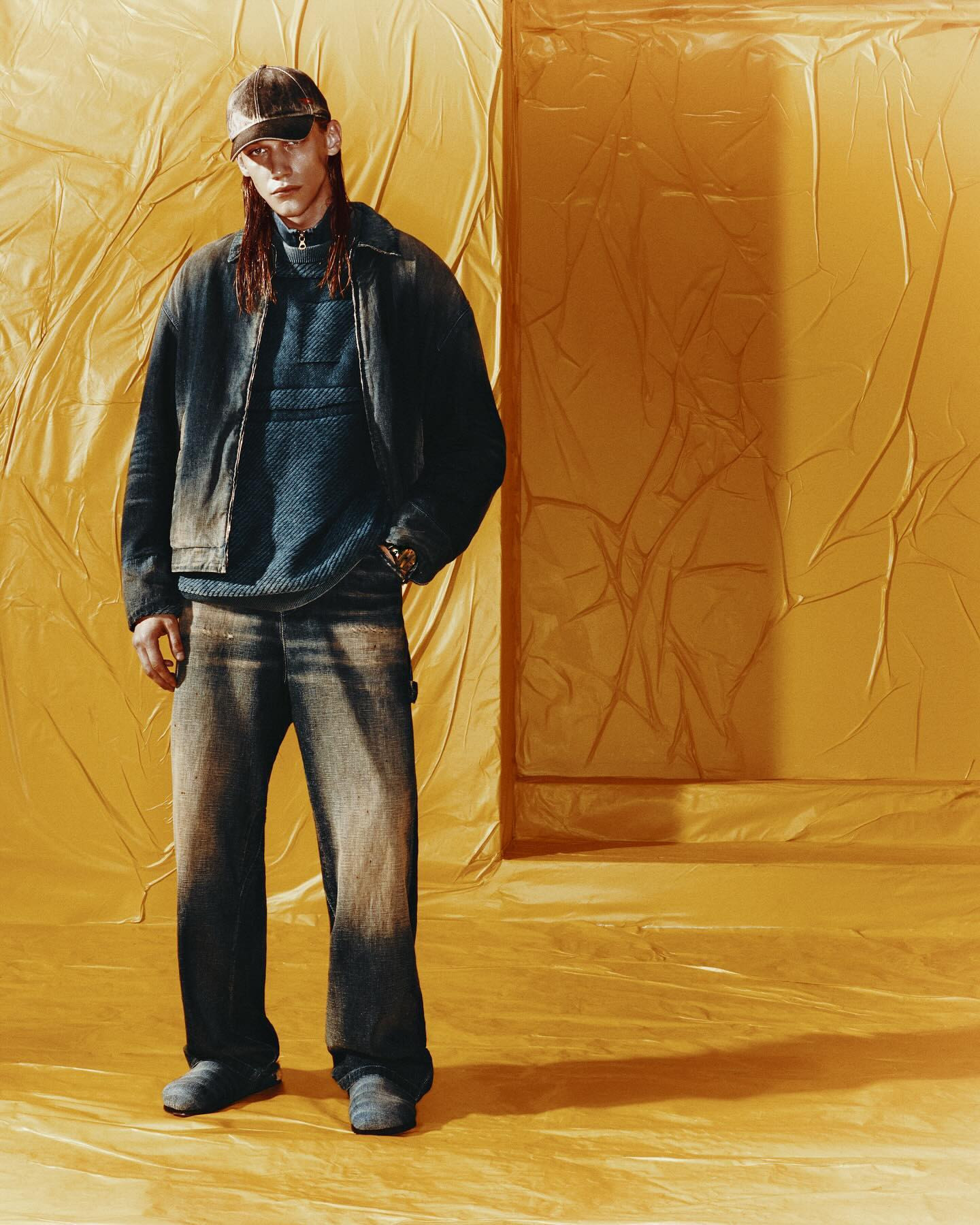 Young adult man in a blue sweater, jacket and jeans, wearing sunglasses and cap. The background is yellow textured material. 