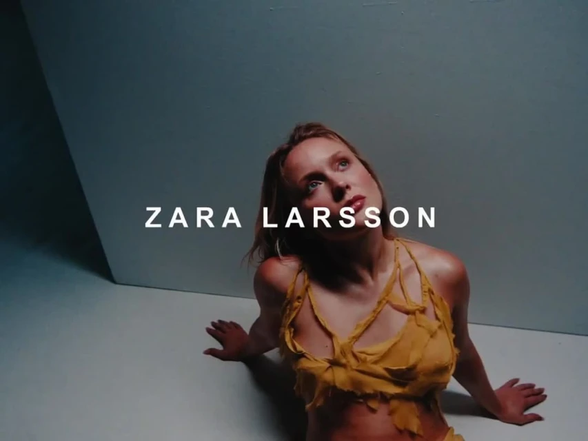 Zara Larsson portrait. A woman in a yellow outfit sits in a photo studio looking up. The backdrop and floor are grey.
