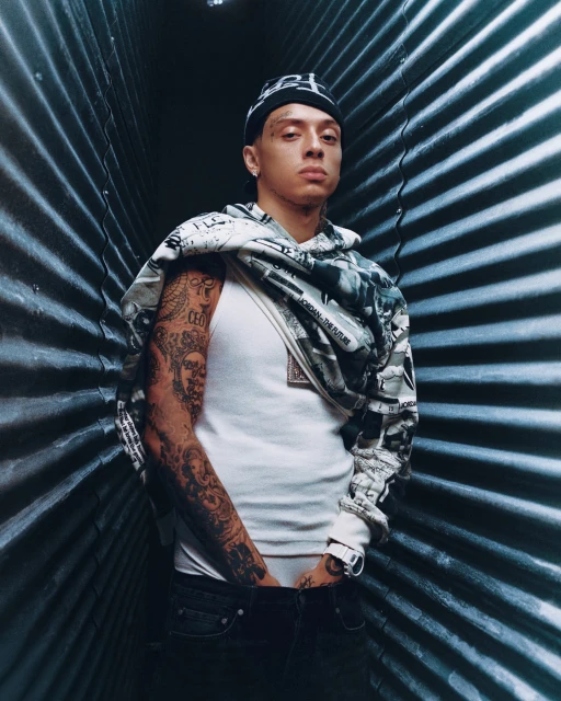 Portrait of a young tattooed man in a white tank top, jeans, and a patterned beanie standing indoors against a corrugated metal wall.