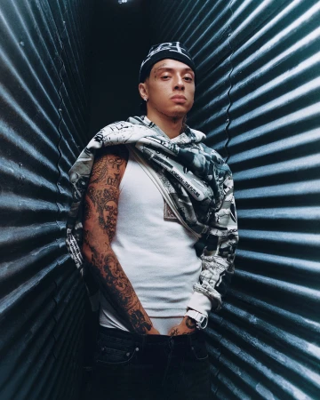 Portrait of a young tattooed man in a white tank top, jeans, and a patterned beanie standing indoors against a corrugated metal wall.
