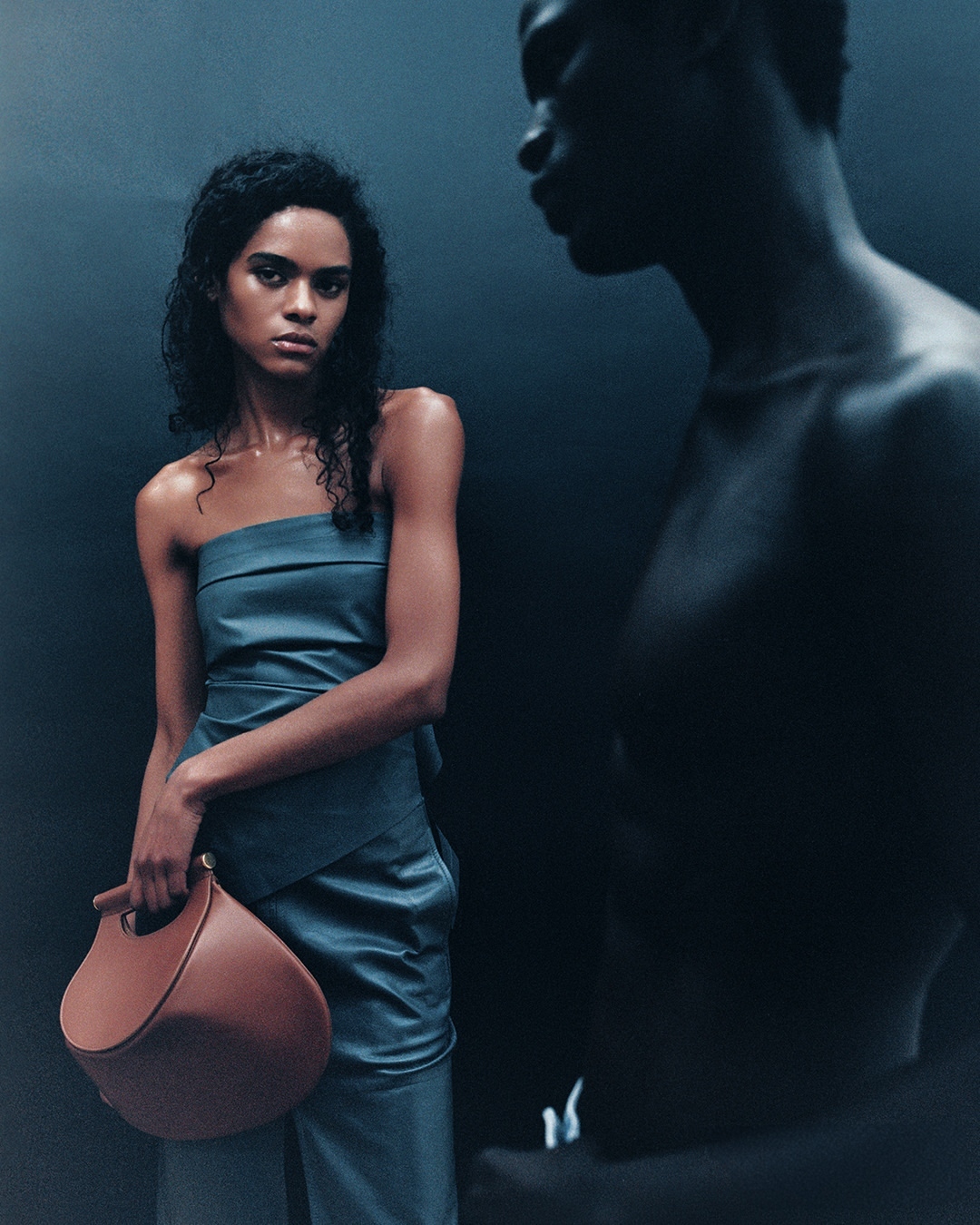 Fashionable woman in a blue one-shoulder dress holding a brown bag with another dark-skinned woman in shadow behind.