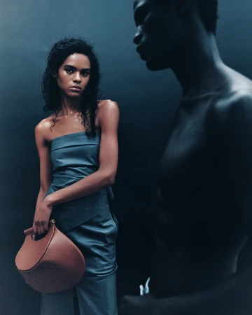 Fashionable woman in a blue one-shoulder dress holding a brown bag with another dark-skinned woman in shadow behind.