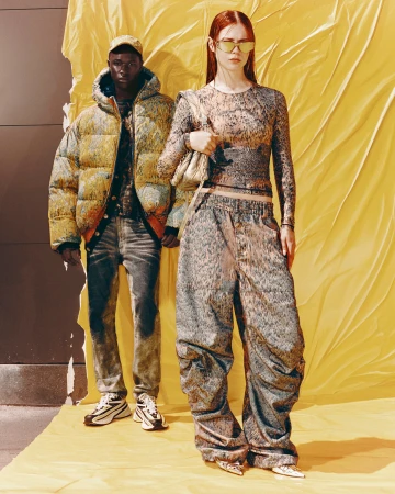 Two models standing against a bright yellow draped background wearing patterned designer streetwear fashion apparel.