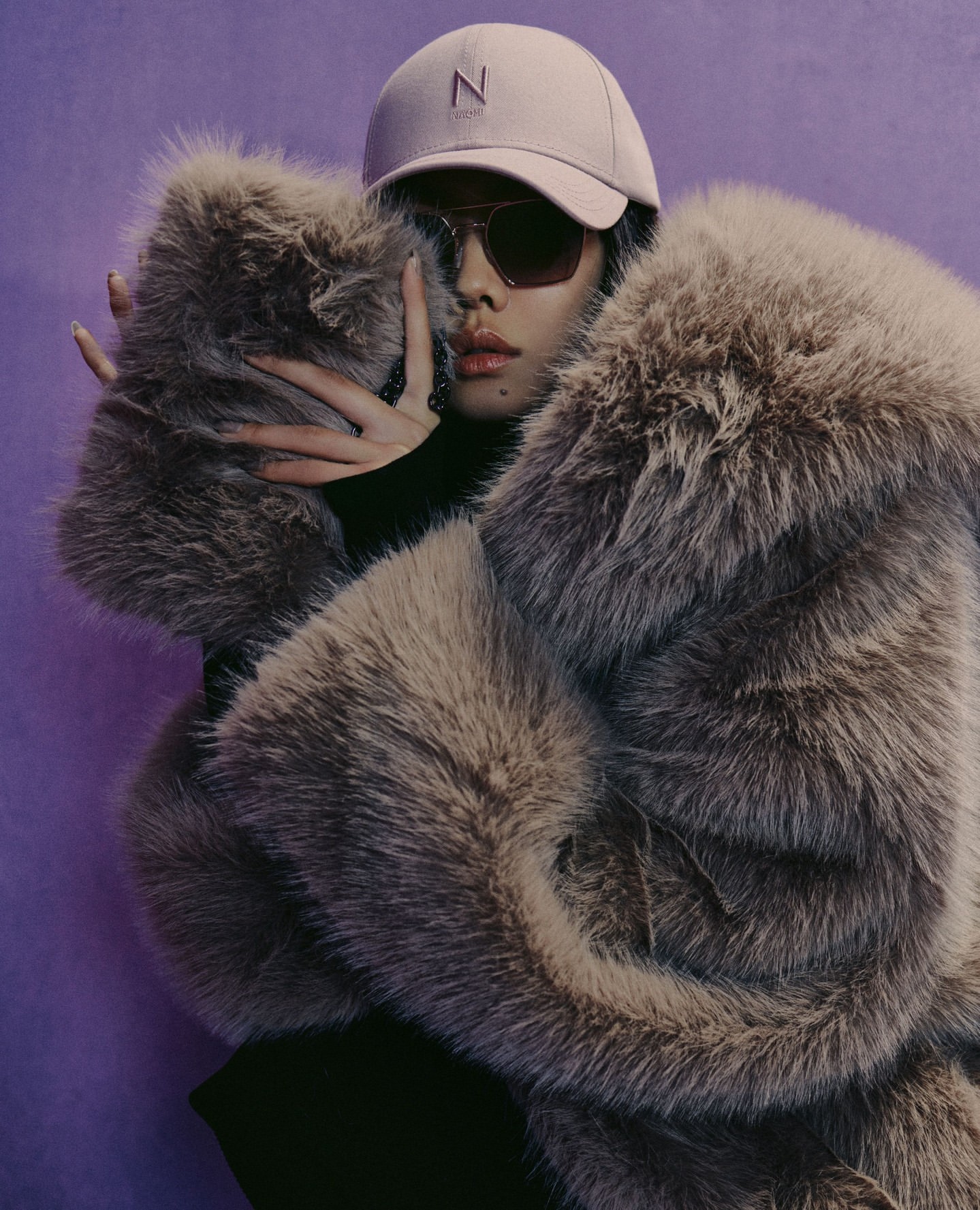 Woman with sunglasses and a cap wearing a large fur coat posing in front of a purple backdrop in a fashion editorial style photograph.