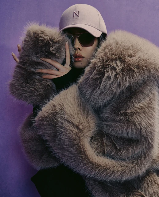 Woman with sunglasses and a cap wearing a large fur coat posing in front of a purple backdrop in a fashion editorial style photograph.