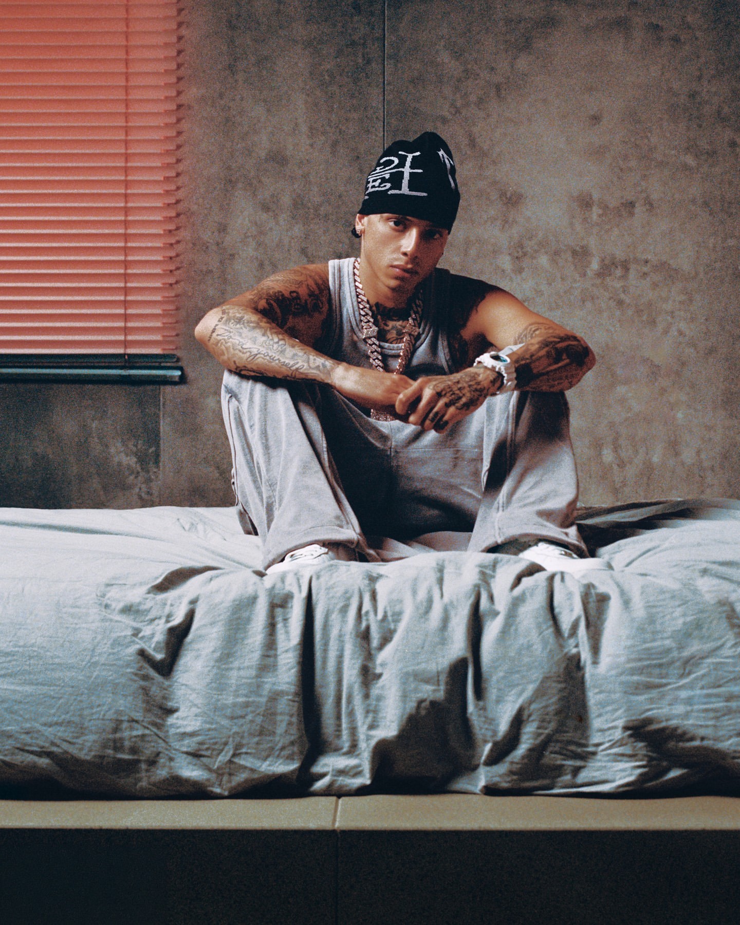 Man in casual streetwear sits on a bed in an indoor setting with a concrete wall, wearing a beanie, tank top, pants and jewelry.