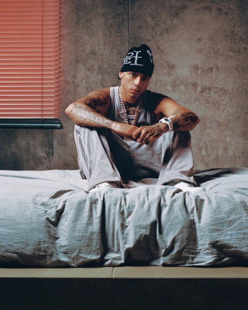 Man in casual streetwear sits on a bed in an indoor setting with a concrete wall, wearing a beanie, tank top, pants and jewelry.