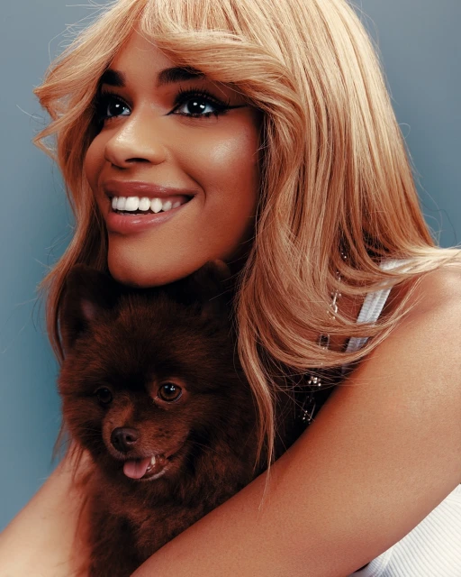 Smiling woman with long blonde wig hugs a brown Pomeranian dog in a close-up portrait against a blue backdrop in a photo studio.
