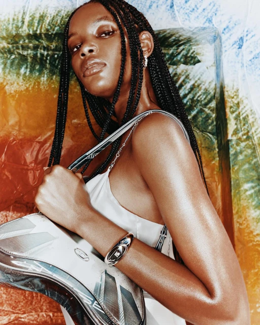 Portrait of a Black woman with braided hair wearing a white dress holding a silver bag in front of a colorful background.