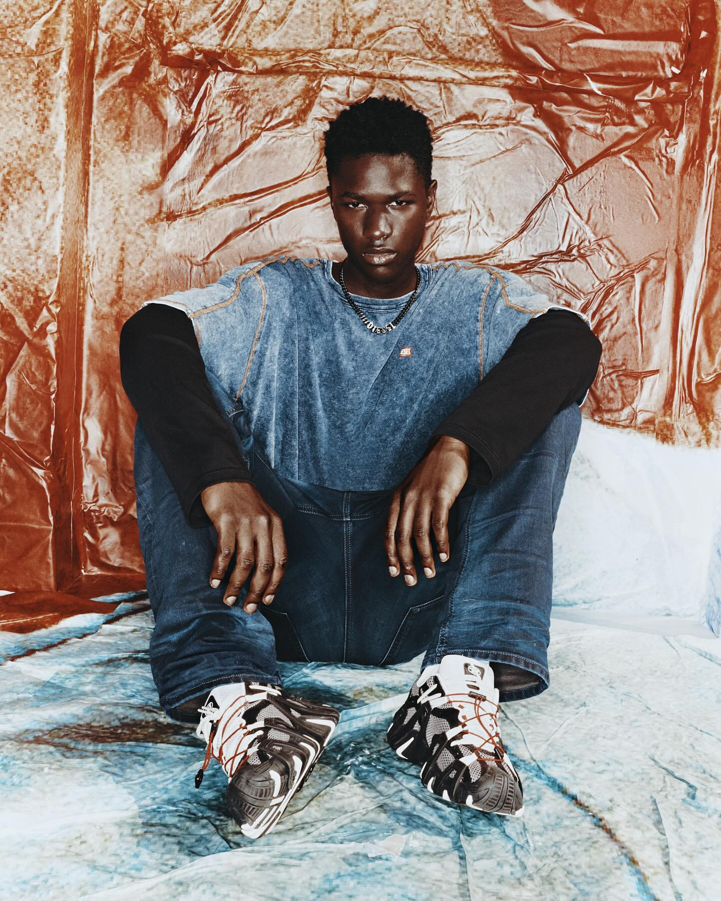 Young man in casual fashion sits against textured backdrop, wearing a blue acid-wash t-shirt, jeans, and chunky sneakers in studio lighting.
