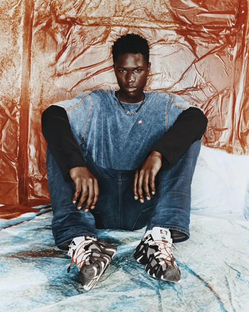 Young man in casual fashion sits against textured backdrop, wearing a blue acid-wash t-shirt, jeans, and chunky sneakers in studio lighting.